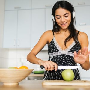 young attractive woman cooking in kitchen in morning, making fruit salad, cutting apple with knife, smiling, happy mood, positive housewife, healthy lifestyle, listening to music on headphones,